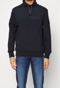 Sudadera negra con cuello alto y cordón ajustable. Presenta un panel de contraste y texto de logotipo en el frente. Tejido suave y ajuste regular.
