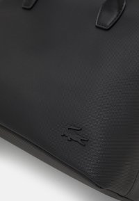 Sac en matériau texturé noir avec des accents en cuir lisse. Présente un logo embossé subtil et des coutures robustes le long des bords.