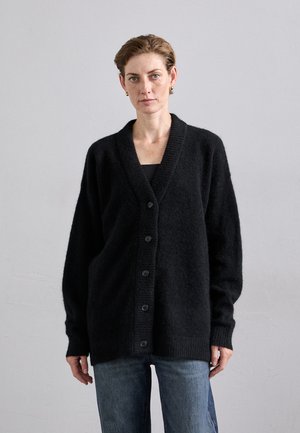 Must missoodev cardigan, mis on valmistatud pehmest ja tekstureeritud kangast. Omab V-kaelust, esipaneeli nuppe ja soonikkoes mansette, kombineeritud siniste teksadega.