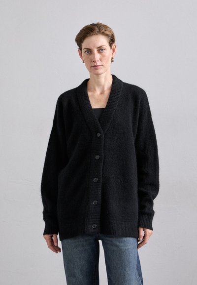 Cardigan noir oversize en tissu doux et texturé. Il présente un col en V, des boutons sur le devant et des poignets côtelés, associé à un jean bleu.