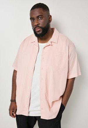 Man met baard die een lichtroze korte mouwen overhemd draagt over een wit shirt en zwarte broek, staand tegen een effen muur.