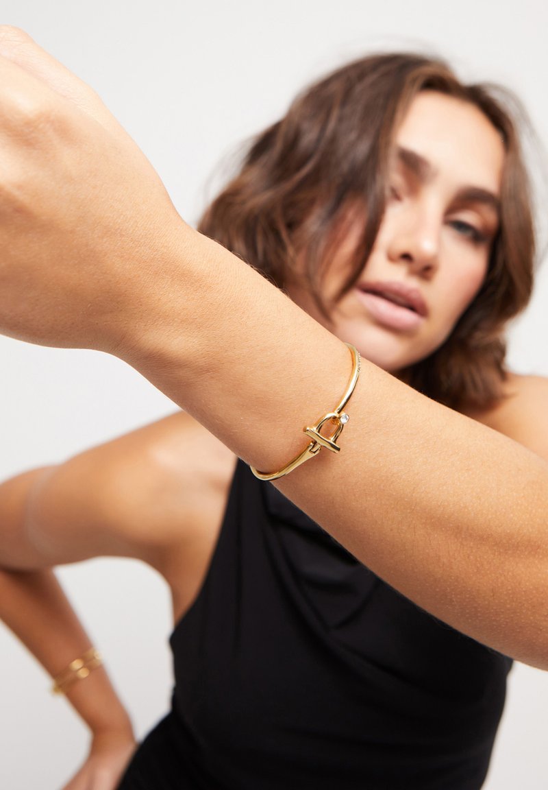 Bracelet en métal doré avec un fermoir décoratif agrémenté d'un petit accent en pierre. Le bracelet est rond et possède une texture lisse.