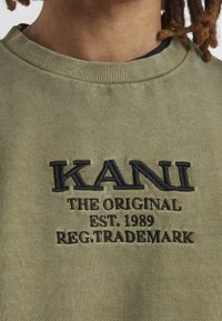 Olivgrön sweatshirt med ribbad halsringning och svart broderad text som lyder "KANI THE ORIGINAL EST. 1989 REG. TRADEMARK."