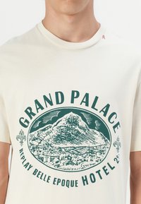 Krémfarget bomulls t-skjorte med grønt grafisk trykk av et fjell og teksten "GRAND PALACE REPLAY BELLE EPOQUE HOTEL." Rund hals.