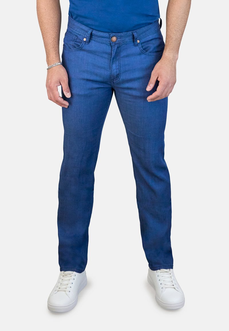 Jeans de hombre en denim azul con un corte recto, cinco bolsillos y detalles en botón de cobre, combinados con zapatillas blancas. Textura suave y lavado medio.
