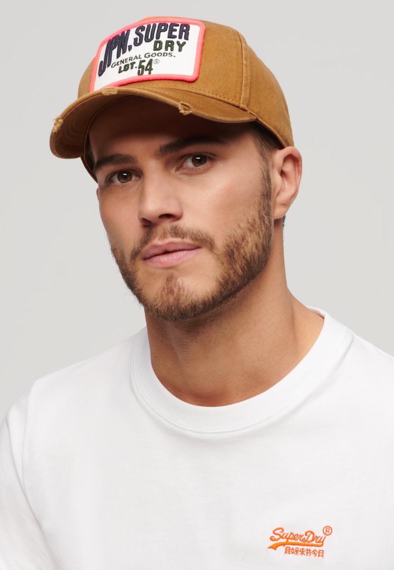Gorra de algodón marrón con un parche rectangular en rosa, rojo y verde que presenta la inscripción "Jon Super Dry General Goods". Se lleva con una camiseta blanca.