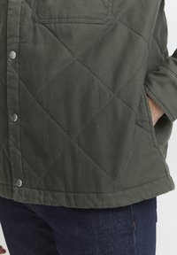 Prošívaná bunda v tmavě zelené látce s diamantovým vzorem, s patentkami a náprsní kapsou. Navržena s uvolněným střihem.