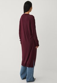 Lange kastanjebruine gebreide cardigan met zijslitsen, geribde manchetten en een ontspannen pasvorm. Gemaakt boven een broek met wijde pijpen van denim. Glad textuur en modern ontwerp.