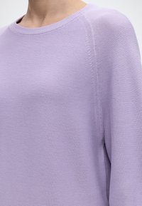 Κοντινό πλάνο ενός ατόμου που φοράει πουλόβερ από υφαντό ύφασμα με ριγέ υφή σε χρώμα λεβάντας, με στρογγυλή λαιμόκοψη και μακριά μανίκια.