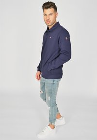 Tummansiniset pitkähihaiset polo paidat, joissa on kaulus ja logopatch, yhdistettynä vaaleansinihin kulutettuihin farkkuihin ja valkoisiin urheilukengiin. Rennon tyylinen asukokonaisuus.