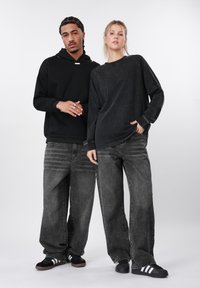 Μαύρο φούτερ με κουκούλα, σκούρο τζιν, άνετη γραμμή. Κορυφές και κάτω μέρος σε αποχρώσεις του γκρι. Μαύρα αθλητικά παπούτσια με λευκές ρίγες. Μινιμαλιστικός σχεδιασμός.