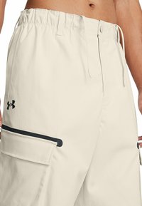 Ljust beige cargoshorts, elastisk midja med dragsko, sidoficka med svart accent, tillverkade av ett strukturerat tyg. Under Armour-logga på vänster sida.