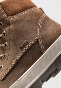 Hnědé semišové polokozačky střední výšky s béžovými šňůrkami, kovovými oky a bílou gumovou podrážkou. Obsahují štítek GORE-TEX a kontrastní prošívání.