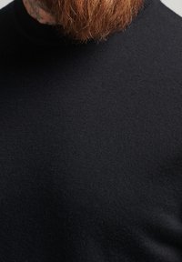 Schwarzer Strickpulloverstoff mit glatter Textur, dessen Nahaufnahme die weiche, gleichmäßige Webart hervorhebt. Rundhals Ausschnitt sichtbar.