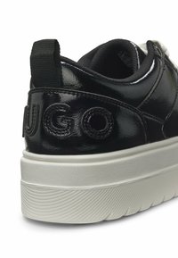 Gros plan sur le talon d'une basket noire brillante avec une languette en tissu noir et une semelle blanche épaisse à rainures, présentant des lettres en relief "UGO".