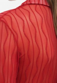 Tissu rouge transparent avec des rayures ondulées verticales couvrant l'épaule et le bras, montrant une légère transparence et des lignes texturées.