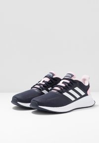 Ett par svarta och rosa Adidas löparskor med vita sulor och tre vita ränder på sidorna, snörda och vinklade åt höger på vit yta.