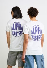 Baltas T-kreklus ar lielu purpura "ALPHA INDUSTRIES" grafiku uz muguras, papildinot ar tekstu "SINCE 1959". Redzama auduma tekstūra.