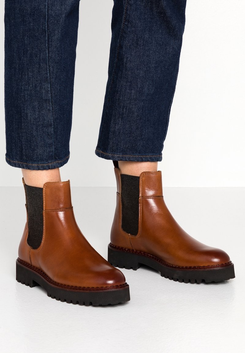 Chevilles portant des bottines Chelsea en cuir marron avec des côtés élastiques noirs et des semelles noires épaisses, associées à un jean en denim bleu foncé.