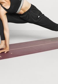 Svart träningskläder inkluderar en crop top med grå ränder och löst sittande byxor. Personen sträcker ut sig på en burgunderröd yogamatta.