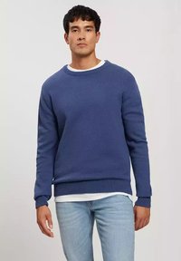 Niebieski sweter dzianinowy z okrągłym dekoltem i ściągaczem na dole. Nosi się go na białej koszuli, ma długie rękawy i luźny krój.