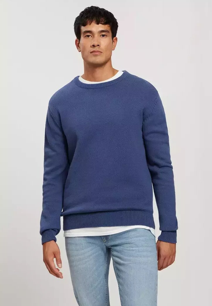 Niebieski sweter dzianinowy z okrągłym dekoltem i ściągaczem na dole. Nosi się go na białej koszuli, ma długie rękawy i luźny krój.