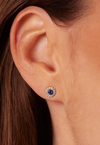 Ronde stud oorbel met een blauwe edelsteen in het midden, omringd door kleine heldere kristallen, gezet in een zilverkleurig metaal, gedragen in het oor.