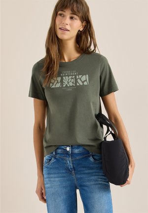 Femme portant un t-shirt graphique couleur olive et un jean bleu, tenant un sac à main noir dans la main gauche, sur un fond uni.