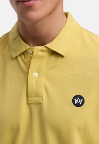 Gul polotröja i texturerad bomull, med en svart rund logo på vänster bröst och en tvåknappsknäppning vid kragen.