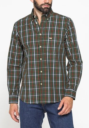 Uomo che indossa una camicia a quadri verde, marrone e bianca con bottoni e maniche lunghe e jeans blu, in piedi su sfondo bianco.
