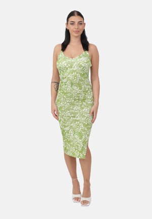 Donna in piedi, indossa un vestito midi verde senza maniche con stampa floreale bianca e sandali bianchi con tacco, su sfondo bianco uniforme.