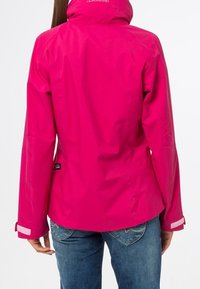 Donna che indossa una giacca impermeabile rosa brillante con cappuccio, in piedi con le spalle rivolte verso la telecamera e le mani rilassate ai lati.