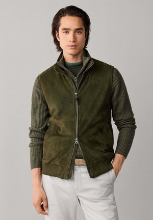 Joven con chaleco verde de ante con cremallera sobre un jersey de punto verde y pantalones de color claro, de pie contra un fondo gris liso.