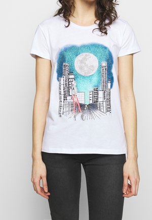 Žena v bílém tričku s grafikou městského panoramatu, velkým měsícem a postavou s dlouhýma růžovýma nohama kráčející po silnici v noci.