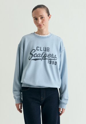 Kobieta w jasnoniebieskiej bluzie czytająca "Club Scalpers 1990" i ciemnych dżinsach, stojąca na jednolitym jasnym tle.