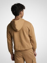 Hanorac cu glugă de culoare camel, cu textură moale, manșete elastice și tiv inferior. Dispune de un buzunar tip cangur și croială casual, asortat cu pantaloni de aceeași culoare.