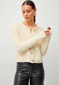 Cardigan duveteux de couleur crème avec un col rond, des boutons dorés et des manches longues, associé à un pantalon noir ajusté. Texture douce et design cropped.