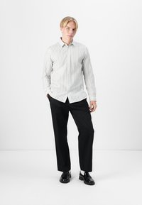 Camicia bianca a maniche lunghe con righe verticali nere, colletto button-down, abbinata a pantaloni neri e scarpe nere. Design minimalista.