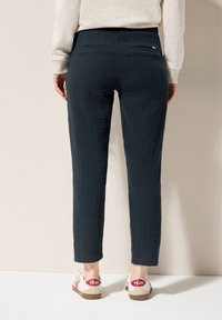 Marineblauwe textuurbroek met een relaxed fit, voorzien van achterzakken en een elastische tailleband, gecombineerd met beige schoenen met rode accenten.