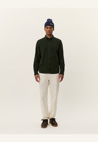 Green corduroy button-up shirt with long sleeves, paired with cream-colored trousers and dark brown shoes. Navy beanie with logo.