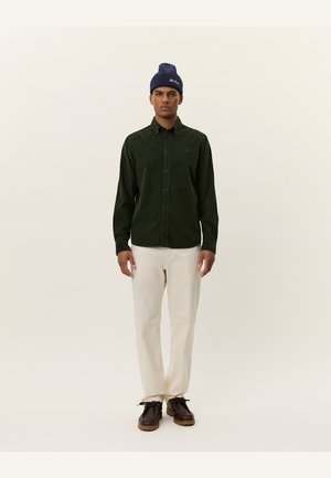 Groene corduroy button-up shirt met lange mouwen, gecombineerd met crèmekleurige broek en donkerbruine schoenen. Marineblauwe beanie met logo.