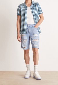 Chemise à manches courtes avec un motif à carreaux bleu et blanc, assortie à un short en denim léger et usé de couleur bleue et des baskets blanches avec des accents de couleur.