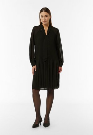 Robe noire plissée avec des manches longues, un col à nouer et une coupe fluide. Portée avec des collants noirs transparents et des chaussures à talons noires.