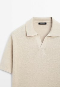 Beige neulepolo, jossa on lyhyet hihat, ribbireunus ja V-pääntie. Ominaisuuksia pehmeä rakenne ja hienovarainen, tasainen kuviointi.