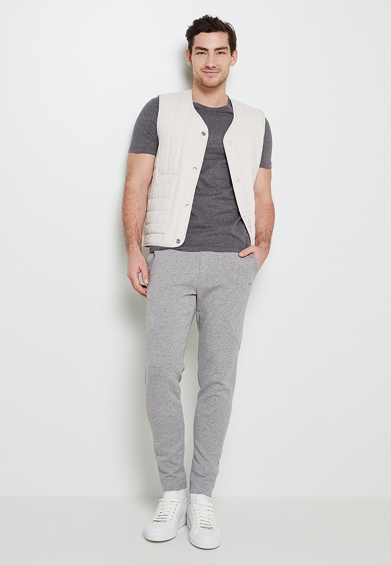 Beżowa pikowana kamizelka na szarym t-shircie, w połączeniu z szarymi dresami i białymi sneakersami. Strój charakteryzuje się prostym, casualowym stylem.