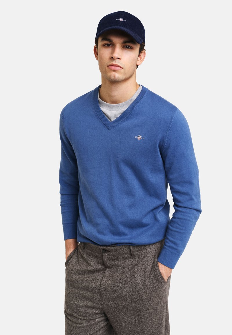 Suéter azul de cuello en V de tejido suave, combinado con pantalones de cuadros grises. Se lleva una camiseta gris debajo y se usa una gorra azul marino.