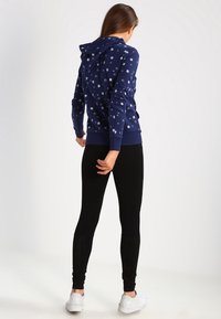 Marinblå hoodie med vitt stjärnmönster, känguruficka och justerbar huva, matchad med svarta leggings och vita sneakers.