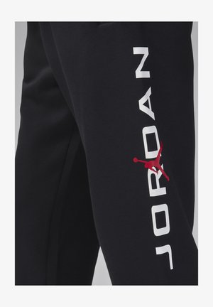 Pantaloni da tuta neri con un logo bianco "JORDAN" lungo la gamba, accentuato da un simbolo Jumpman rosso. Realizzati in morbido tessuto testurizzato.