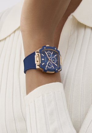 Blue watch with gold accents, square face, three subdials, and rubber strap, worn on a wrist, paired with a ribbed white sweater.