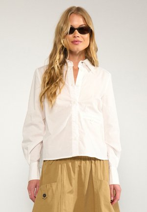 Femme aux longs cheveux blonds portant des lunettes de soleil noires, une chemise blanche boutonnée et une jupe beige avec de grandes poches.